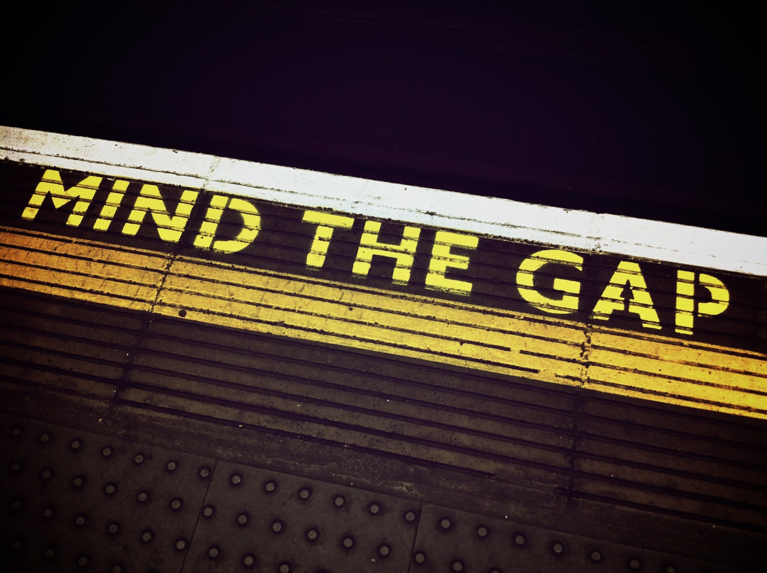 Yellow text on train platform edge reads 'MIND THE GAP' with tactile paving below.