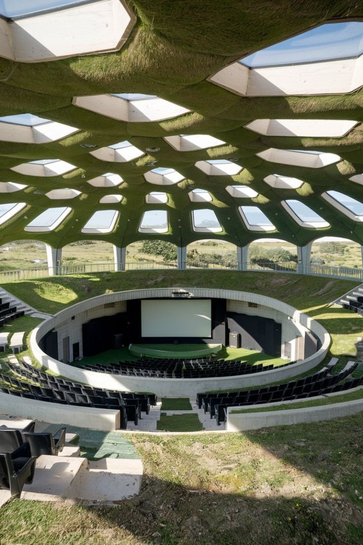 Open-air amphitheater with circular seating, grass-covered roof, and large projector screen.