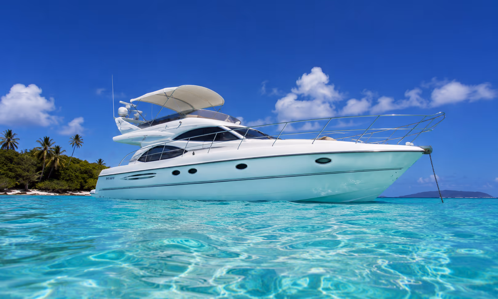Yacht blanc ancré sur une eau turquoise claire près d'une île avec des palmiers et ciel bleu.