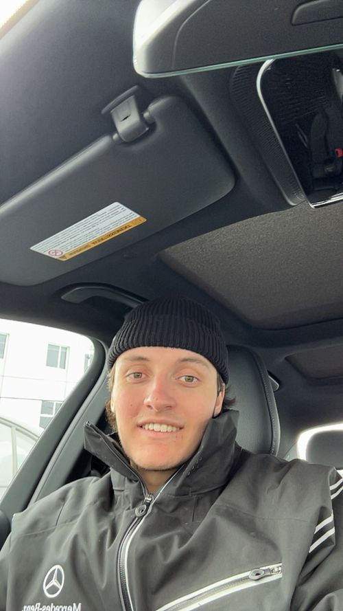 Jeune homme souriant avec un bonnet noir portant une veste Mercedes-Benz à l'intérieur d'une voiture.