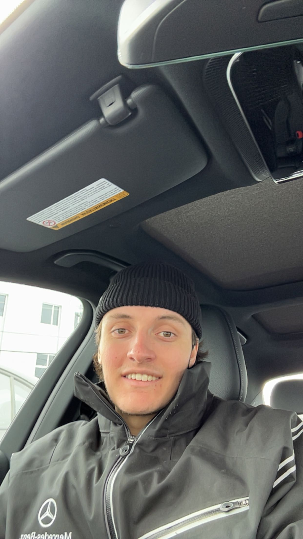 Jeune homme souriant avec un bonnet noir portant une veste Mercedes-Benz à l'intérieur d'une voiture.