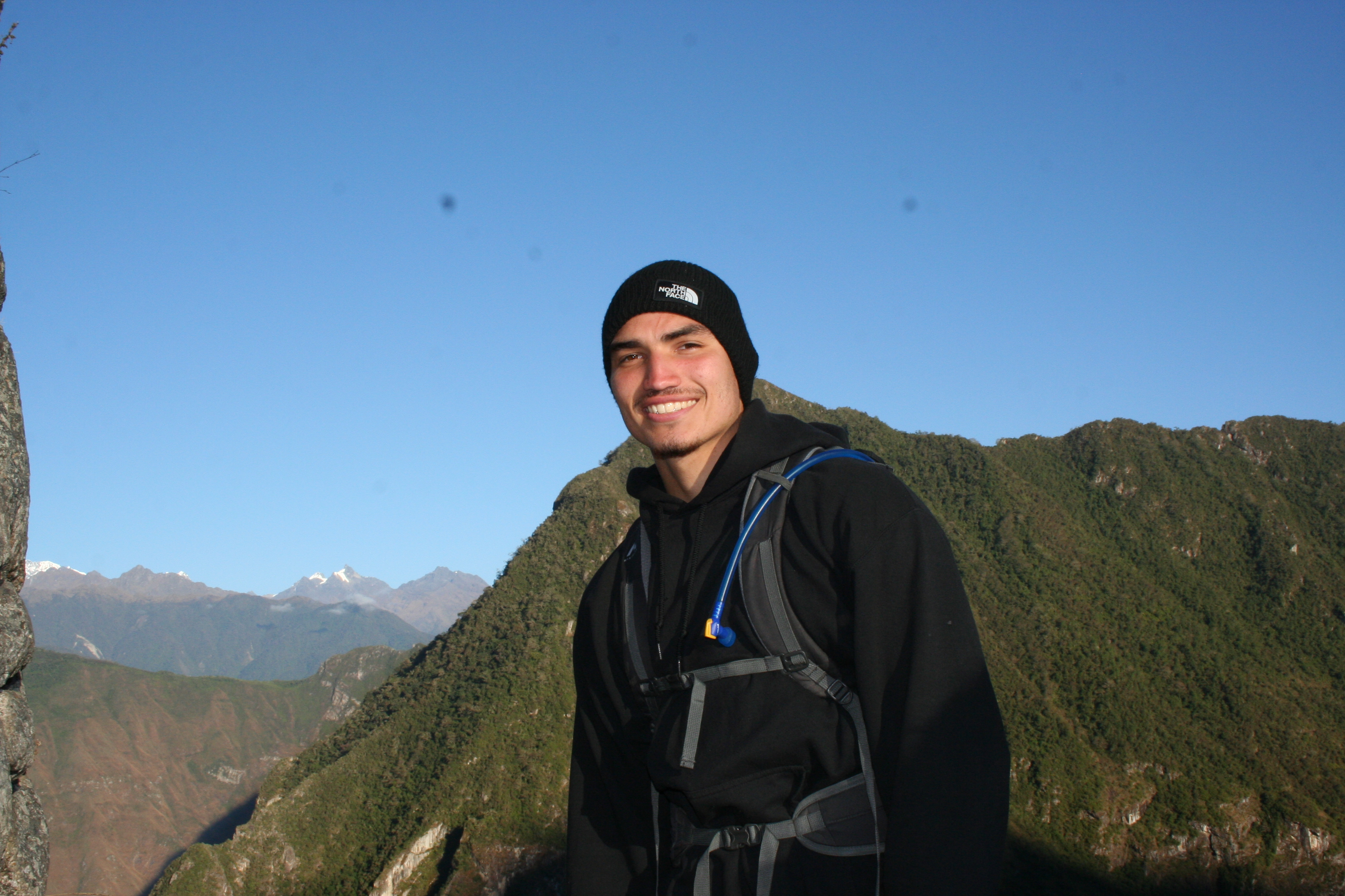 Jeune homme souriant en tenue de randonnée devant une montagne verte sous ciel bleu dégagé.