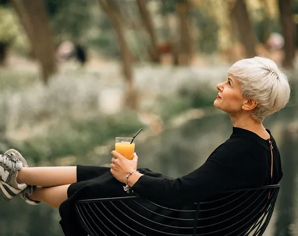 Woman with short gray hair relaxing outdoors holding a glass of orange juice with a straw.