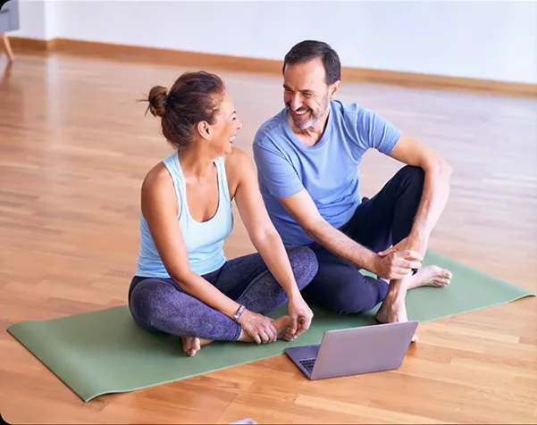 A smiling couple sitting cross-legged on a yoga mat with a laptop on a wooden floor.