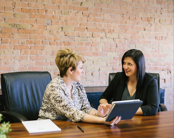 Two women in an office discussing while one shows information on a tablet at a wooden table.