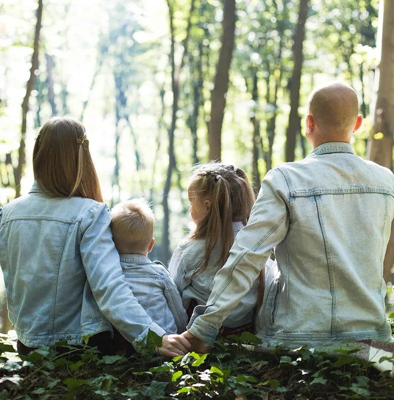 Family of four in denim jackets holding hands sitting outdoors among greenery facing forest trees.