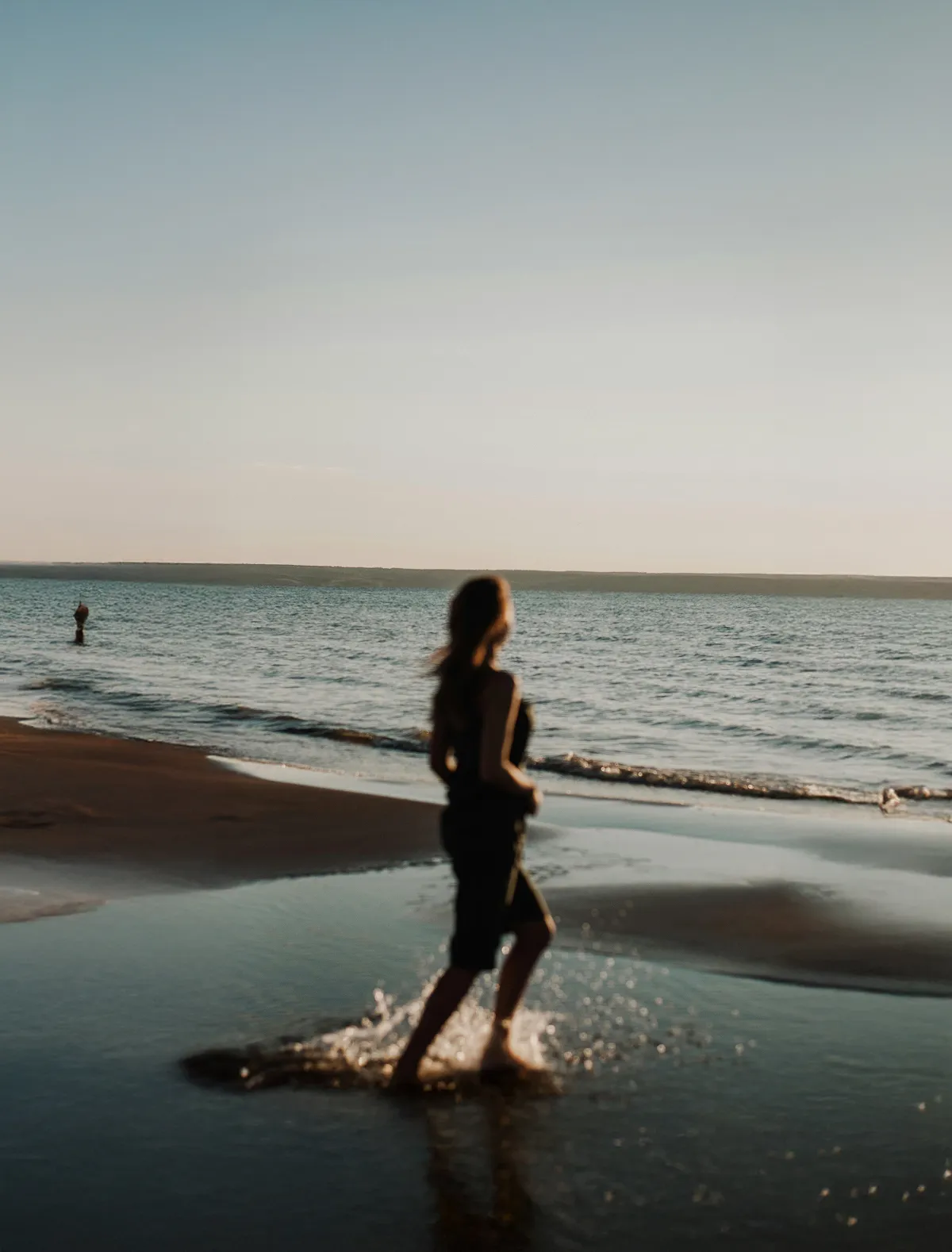 Blurry silhouette of a person walking barefoot splashing water along a shore at sunset.