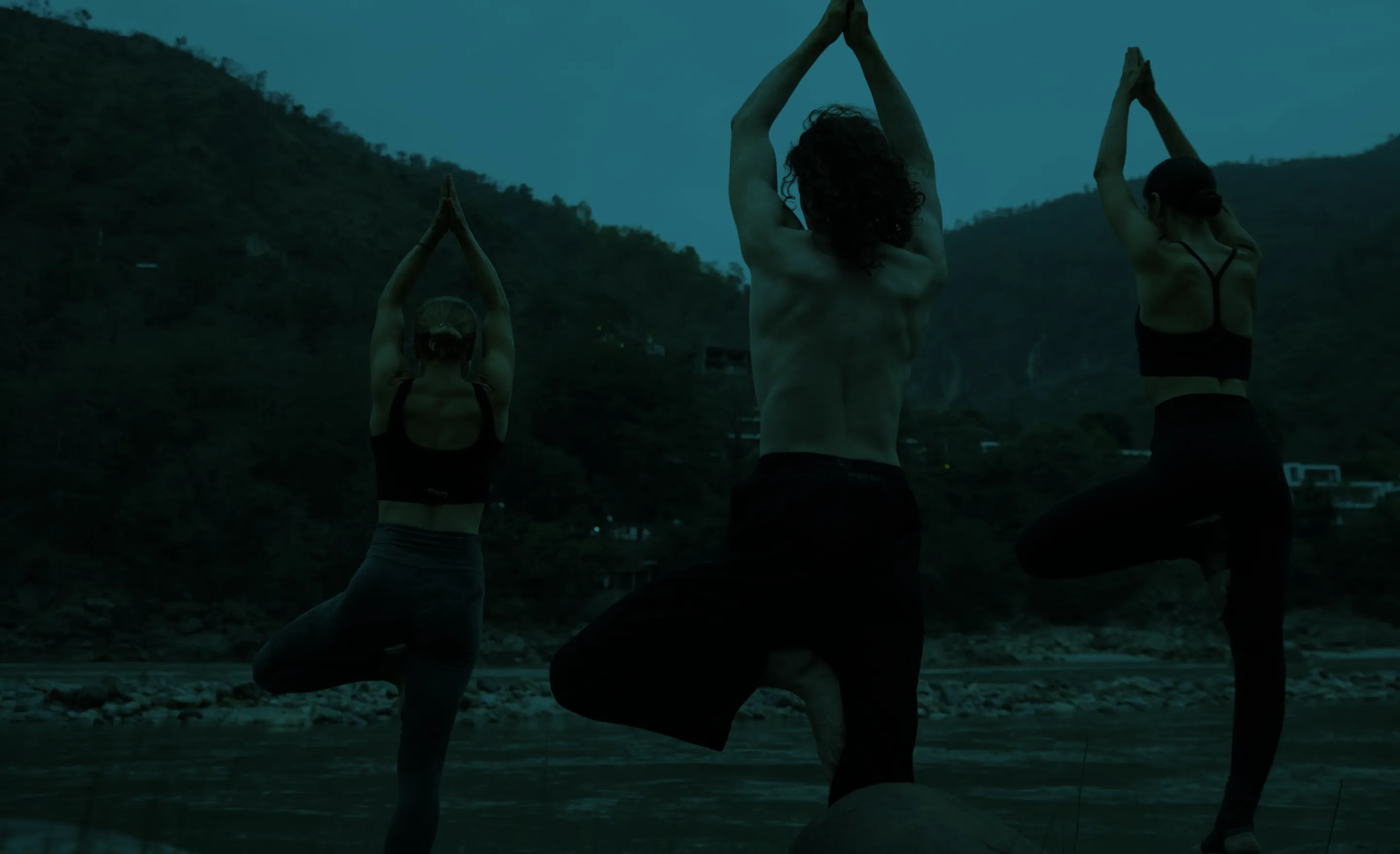 Three people practicing yoga in tree pose near a river with mountain background at dusk.