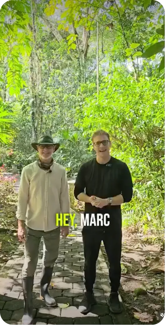 Two men standing on a stone path surrounded by lush green trees and plants outdoors.