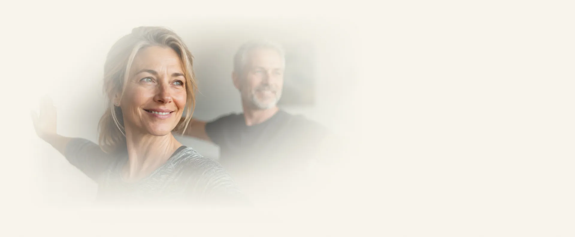Smiling middle-aged woman and man stretching arms in a bright, softly blurred setting.