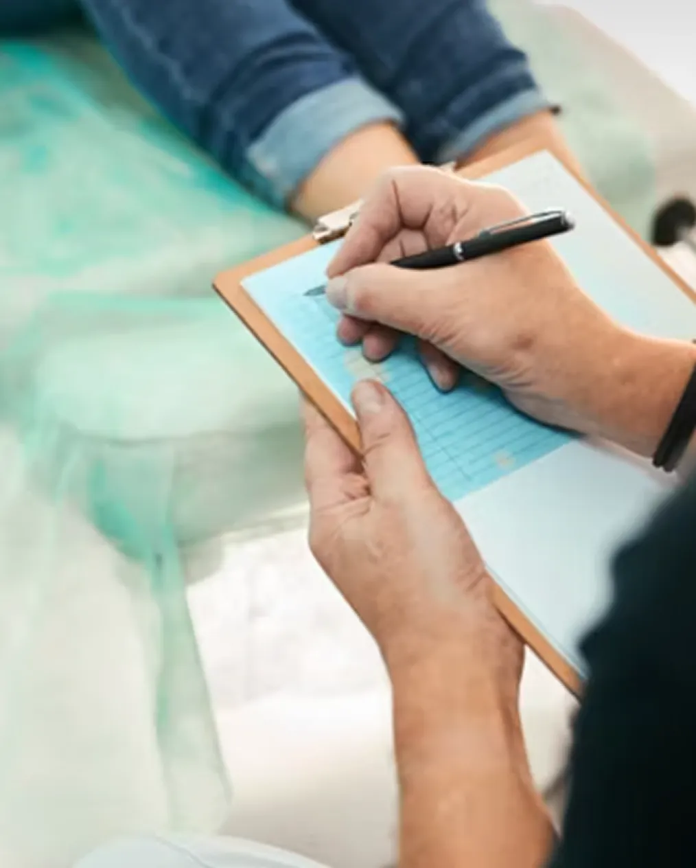Person writing on a blue paper attached to a clipboard near someone sitting with crossed legs.