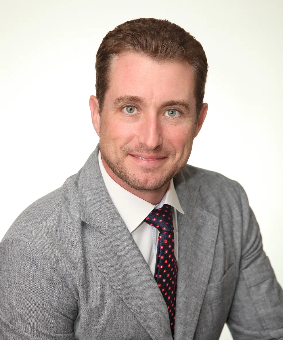 Smiling man with short brown hair in gray suit, white shirt, and patterned tie on a white background.