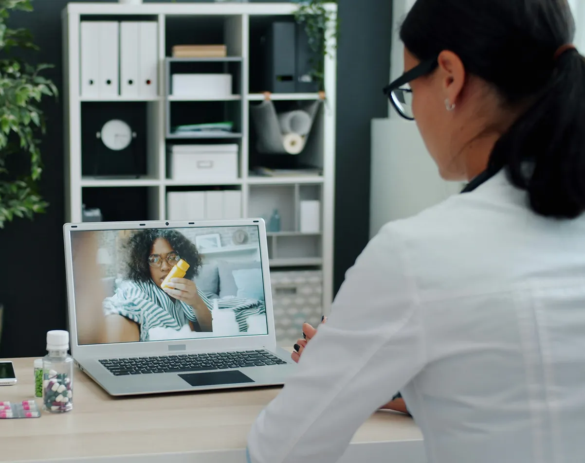 Doctor consulting a patient via video call, patient holds a pill bottle, medicine on desk.
