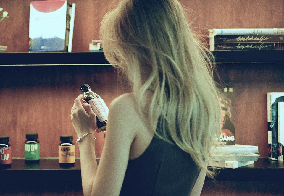 Blonde woman with bracelet examines bottle label in a room with shelves of books and jars.
