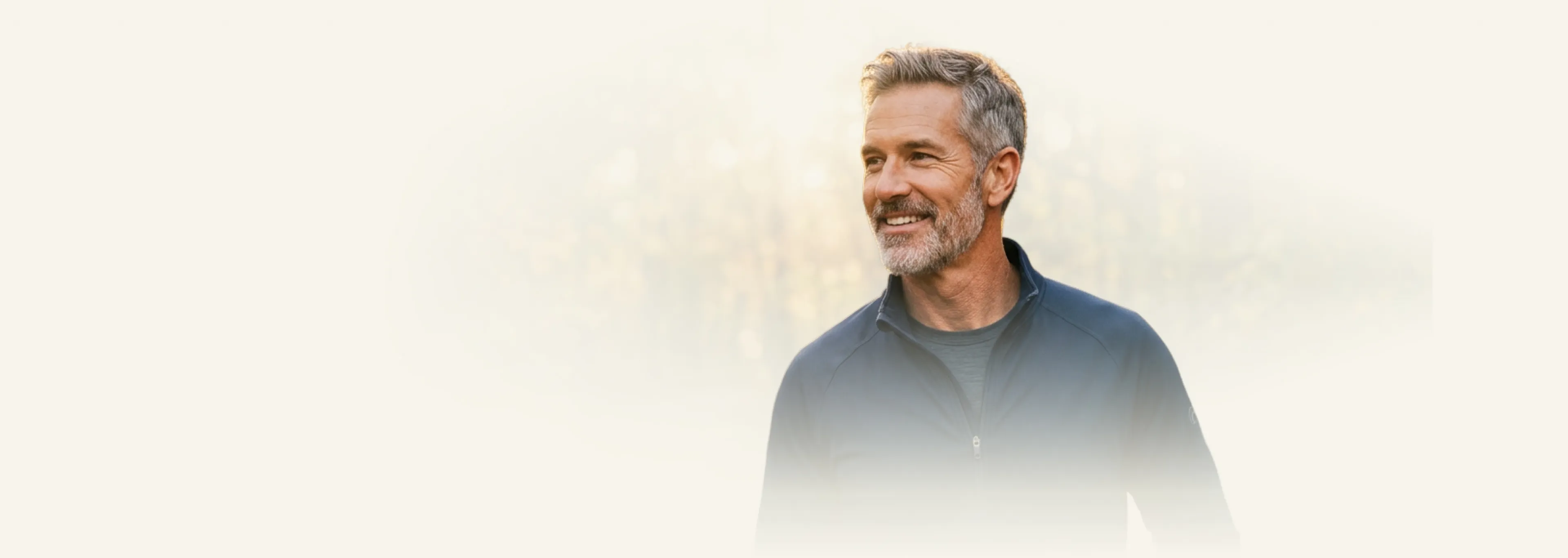 Smiling gray-haired man with beard wearing a dark zip-up jacket looking right on light background.