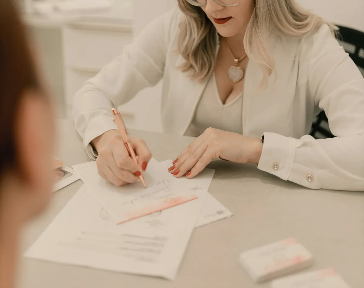 Woman with glasses and red nails signing documents on a desk.