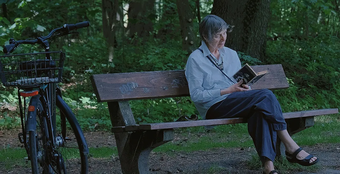 Older woman sitting on a park bench reading a book beside a bicycle in a green wooded area.