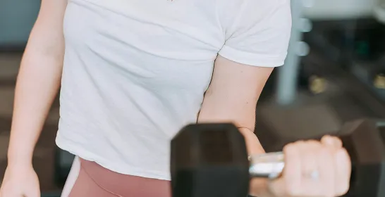 Person lifting a dumbbell with one arm wearing a white top and pink leggings indoors.