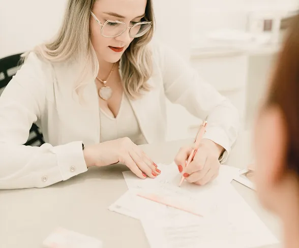Woman wearing glasses writing on paper with a pen at a desk, viewed from an over-the-shoulder perspective.