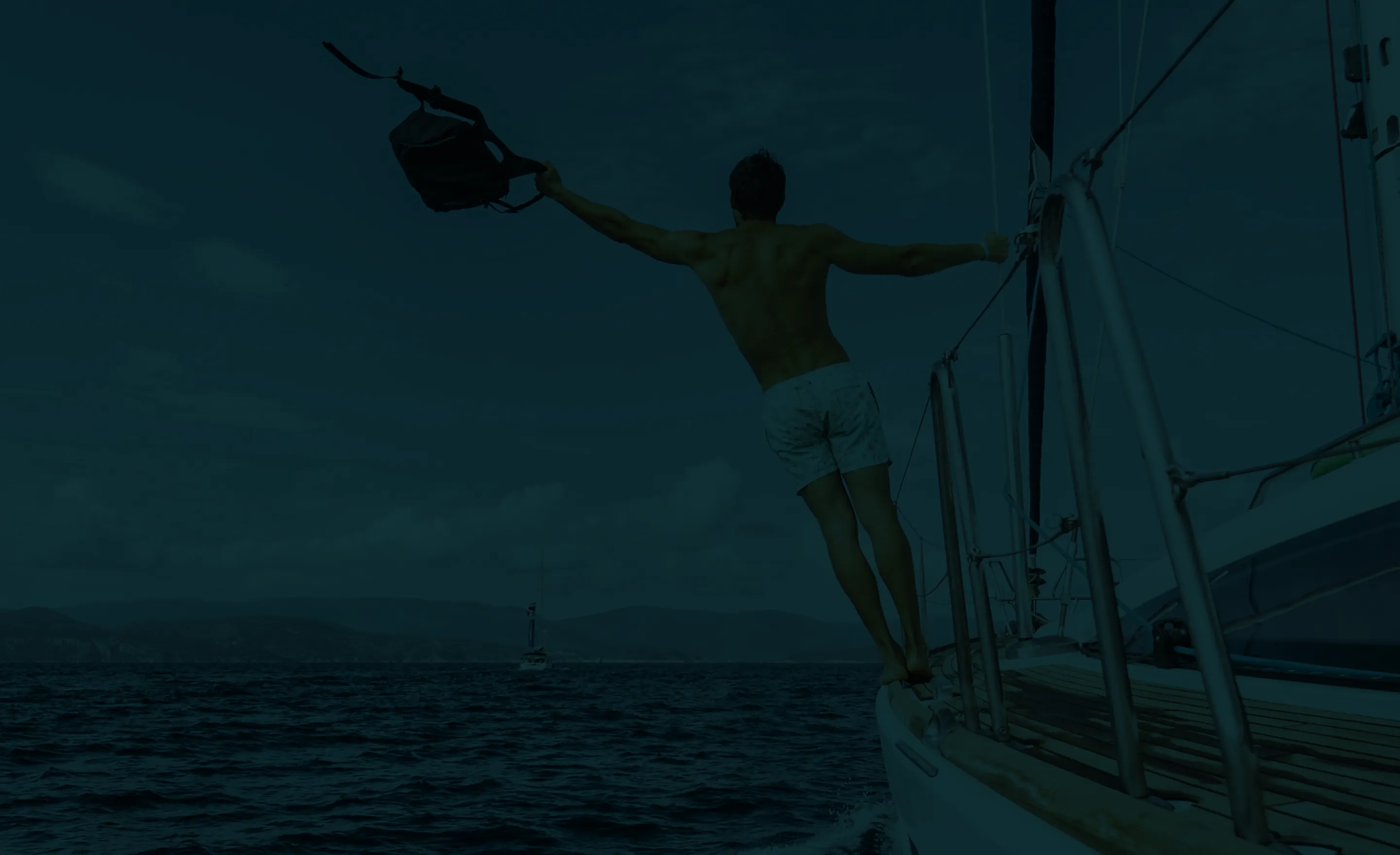 Man in swim trunks stands on boat edge holding a bag out over the ocean.