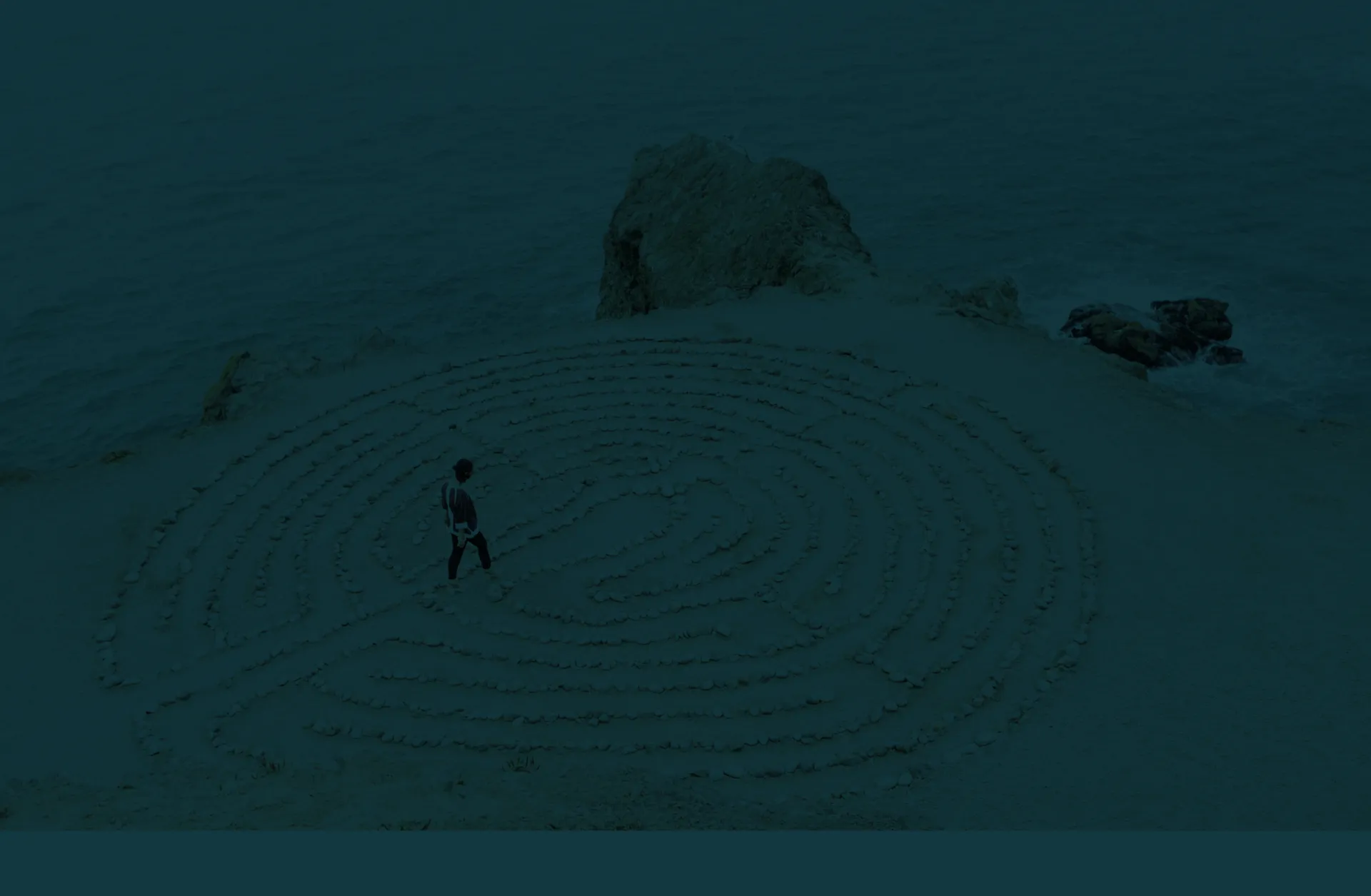 Person walking in a large stone labyrinth on a sandy cliffside above the ocean.