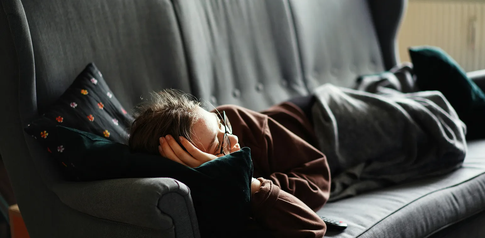 Person lying on a gray couch covered with a blanket, resting their head on pillows with glasses on.