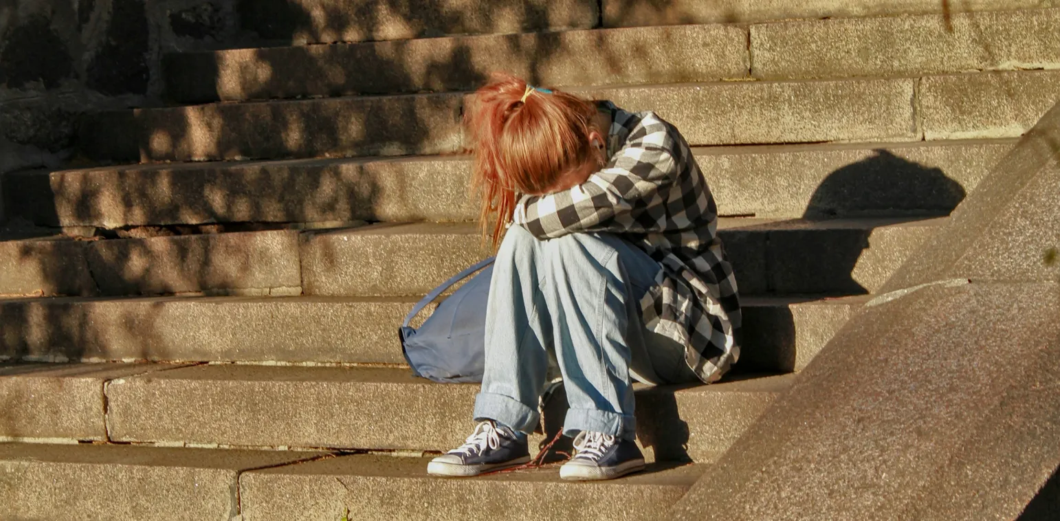 Person in plaid shirt and jeans sitting on stone steps with head resting on arms, appearing sad.