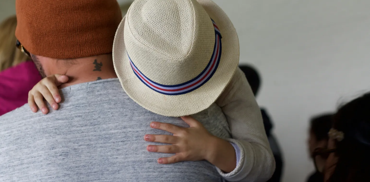 Child wearing a hat hugging an adult from behind, with the adult wearing a brown beanie and gray shirt.