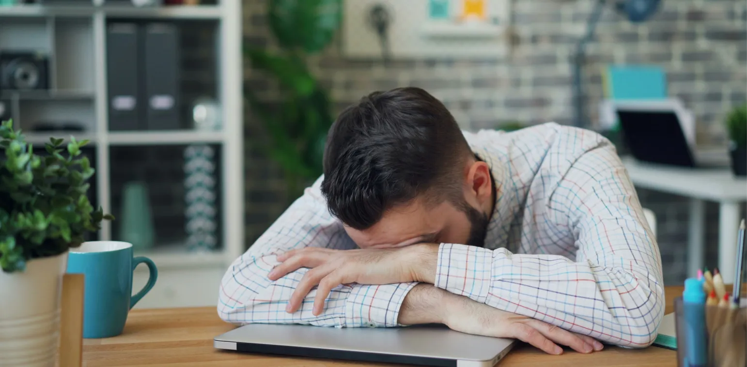 Man with head resting on folded arms on closed laptop, appearing tired or frustrated at desk.