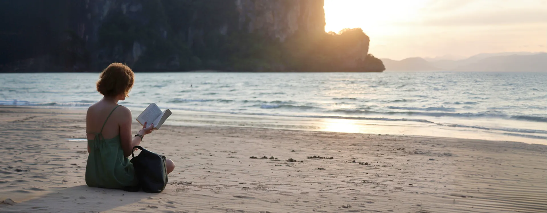 Person in green dress sits on a sandy beach reading a book during sunset near cliffs and sea.