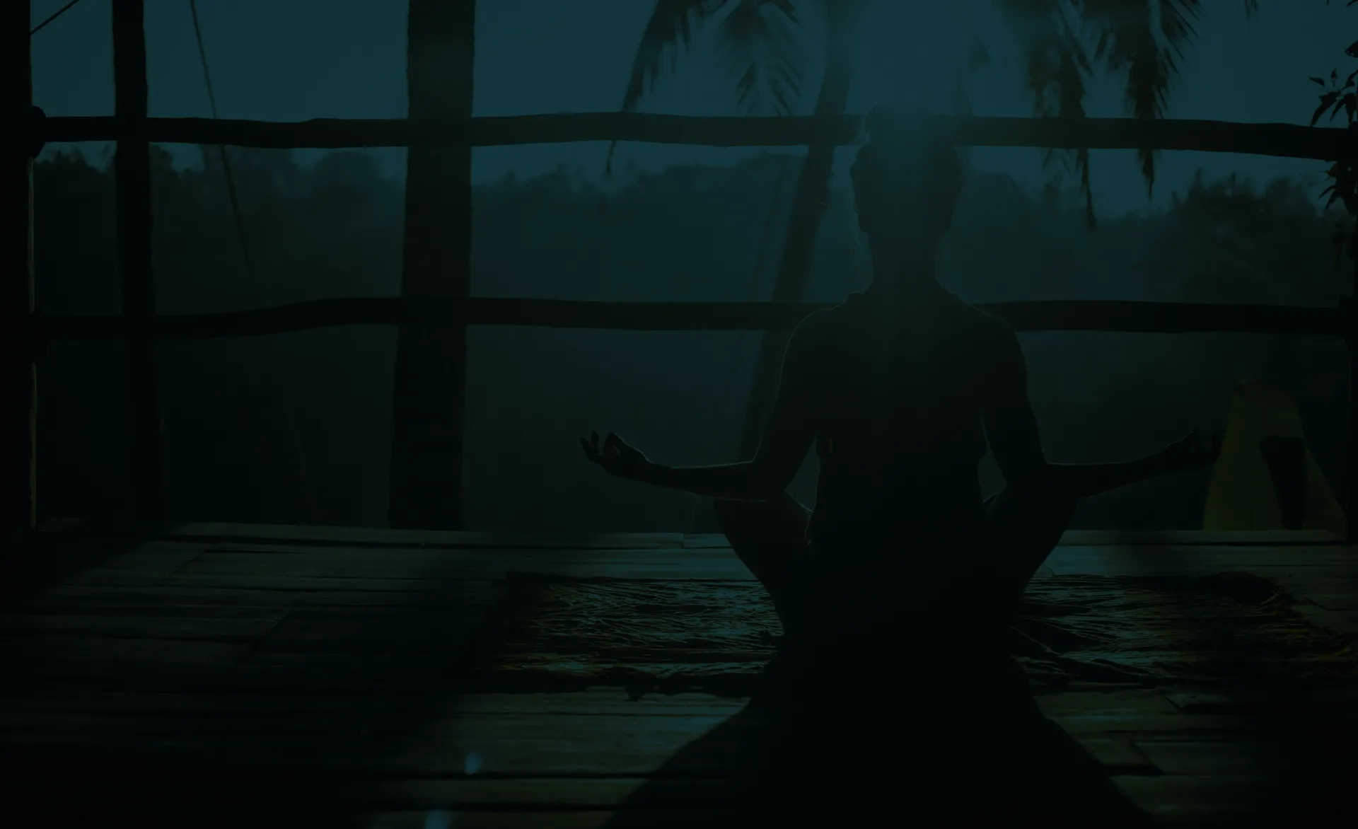 Silhouette of person meditating cross-legged on wooden floor by railing at dusk.