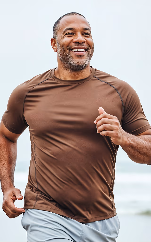 Smiling man wearing brown shirt jogging outdoors near a body of water.