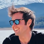 Smiling man wearing sunglasses with reflection of mountains and lake, outdoors with mountains in background.