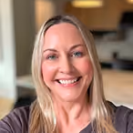 Smiling woman with long blonde hair in a brown top, indoor room background.