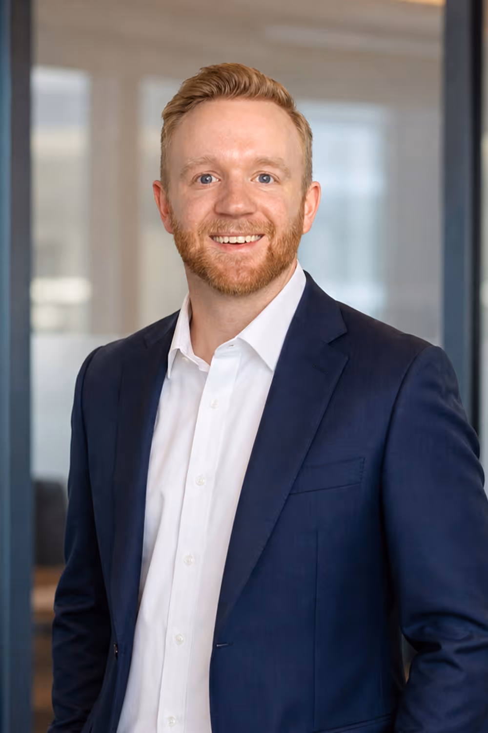 Smiling man with red hair and beard wearing a navy suit jacket and white shirt in an office.