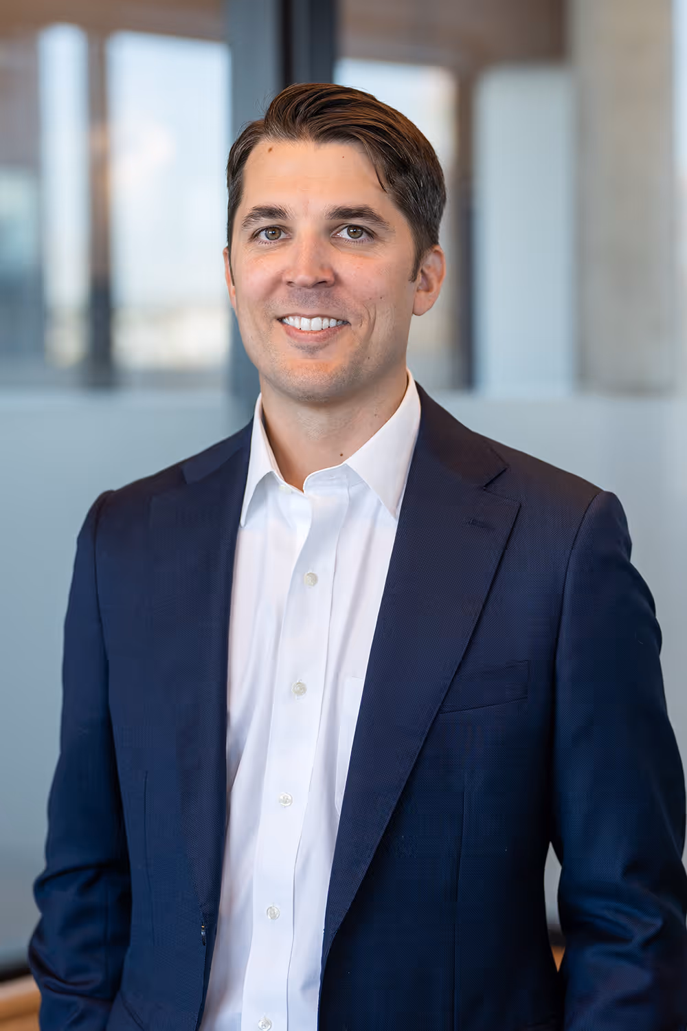 Man in navy suit jacket and white shirt smiling with blurred office background.
