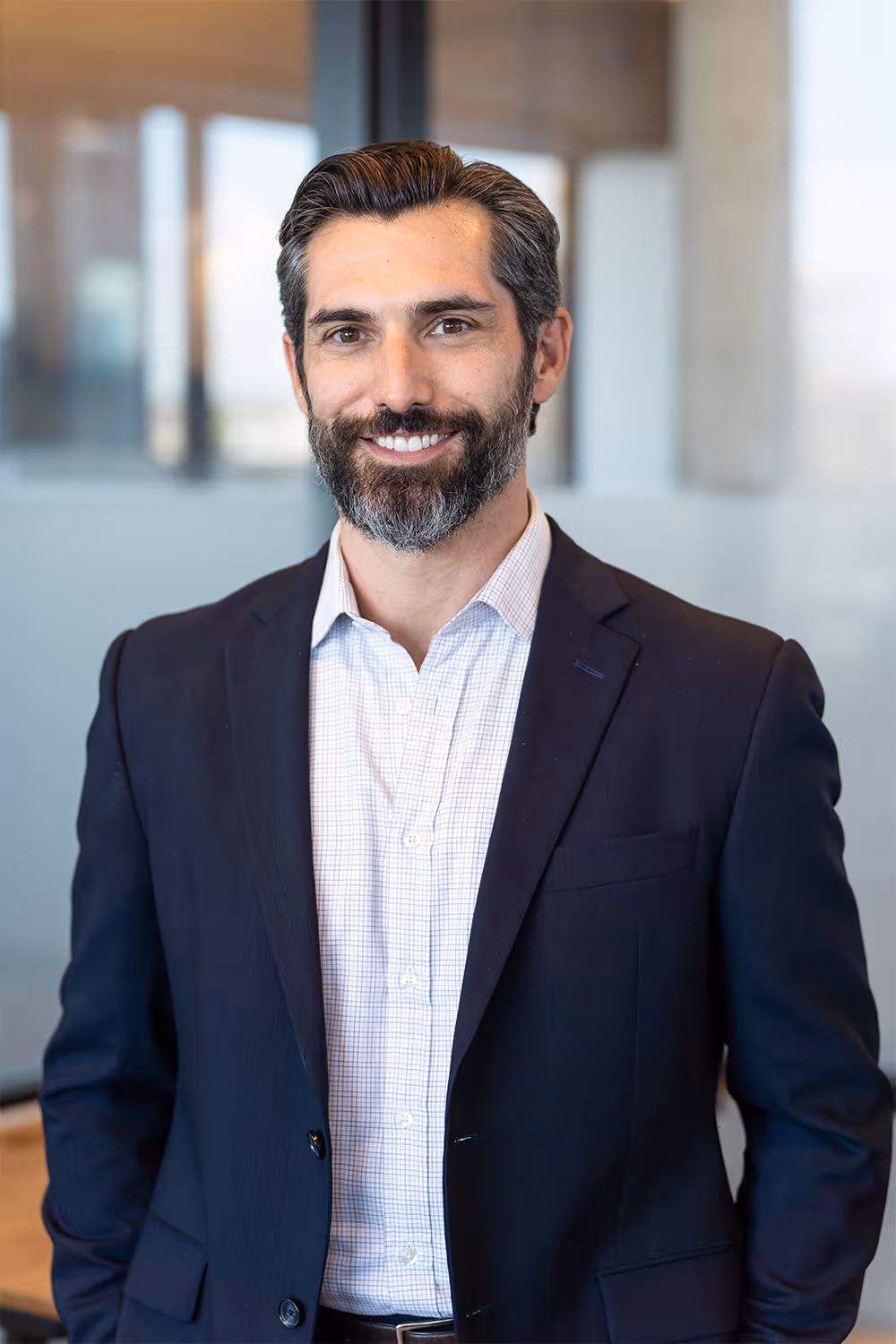 Smiling man with dark hair and beard in white shirt and dark blazer in office setting.