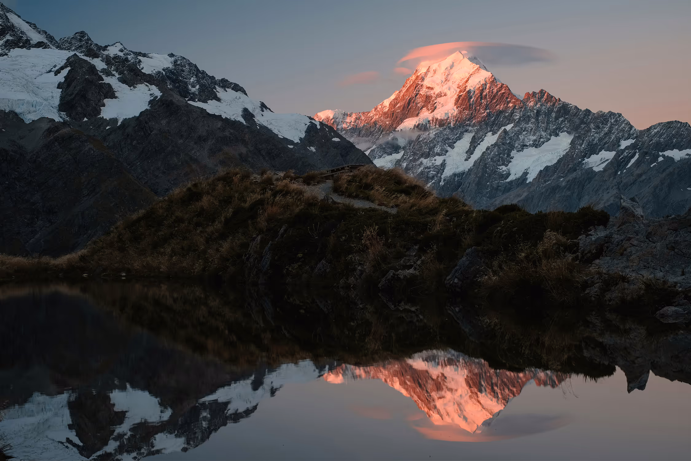 Snow-capped mountain peak illuminated by sunset reflected in a calm lake below.