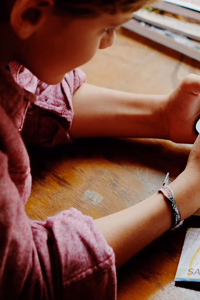 Child in a red shirt using a smartphone at a wooden table near a window.