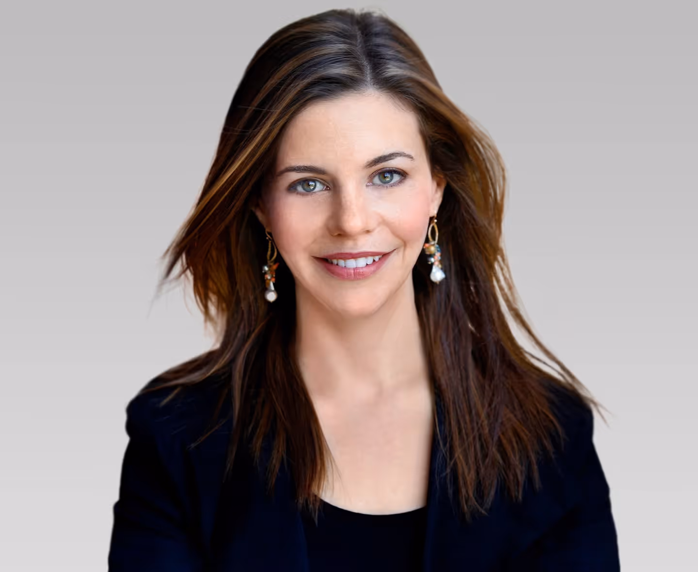 Smiling woman with brown hair wearing dangling earrings and a black top against gray background.
