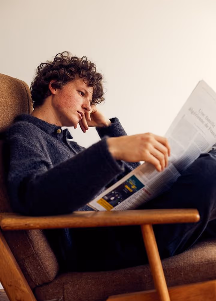 Young man with curly hair sitting in a chair, reading a newspaper thoughtfully.