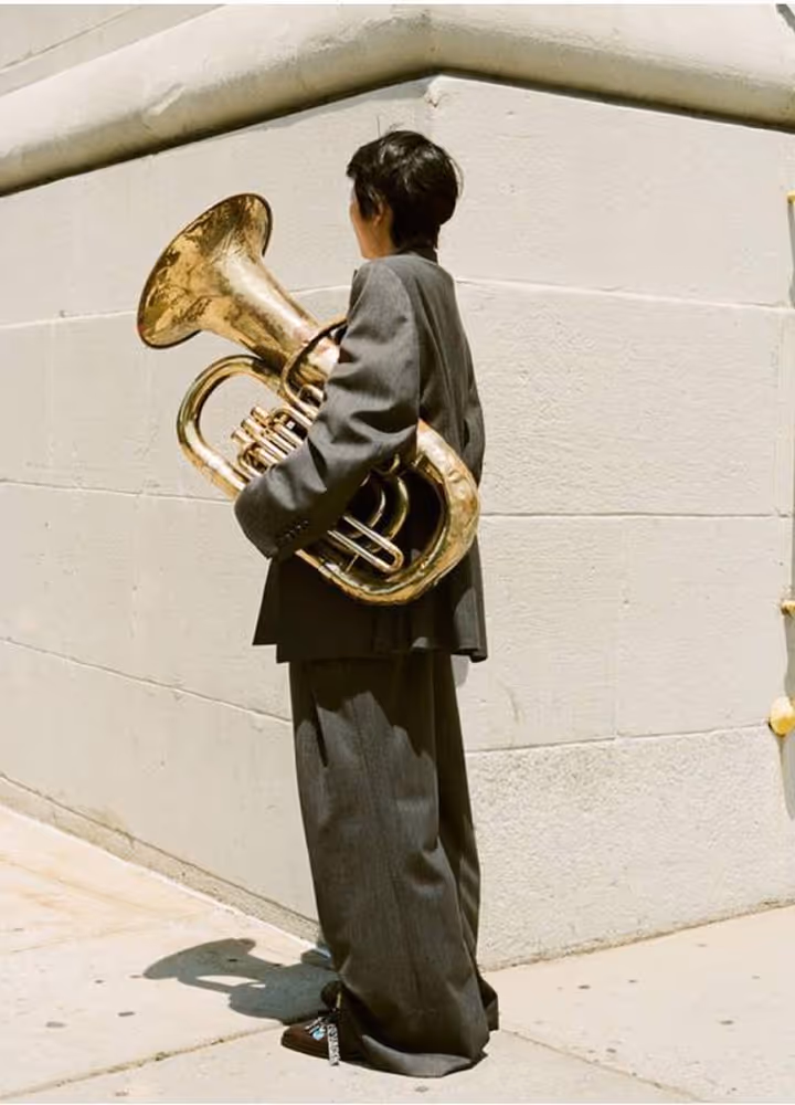 Person in dark oversized suit holding a large brass tuba near a light-colored building corner.