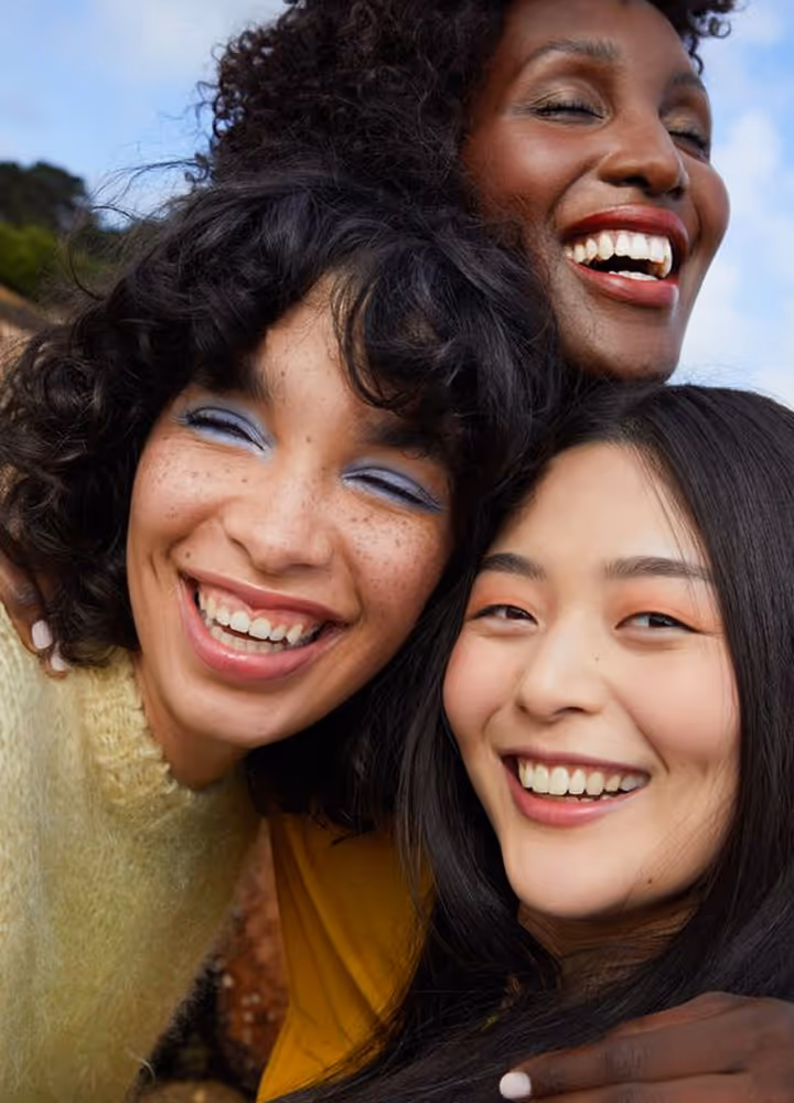 Three diverse women smiling closely together outdoors, showing joy and friendship.