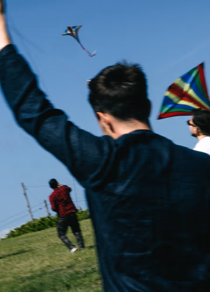 Three people flying colorful kites on a grassy hill under a clear blue sky.