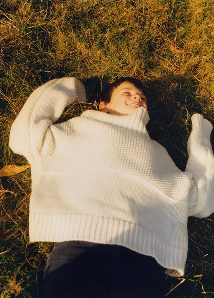 Smiling person lying on grass wearing a large white knit sweater with arms spread out.