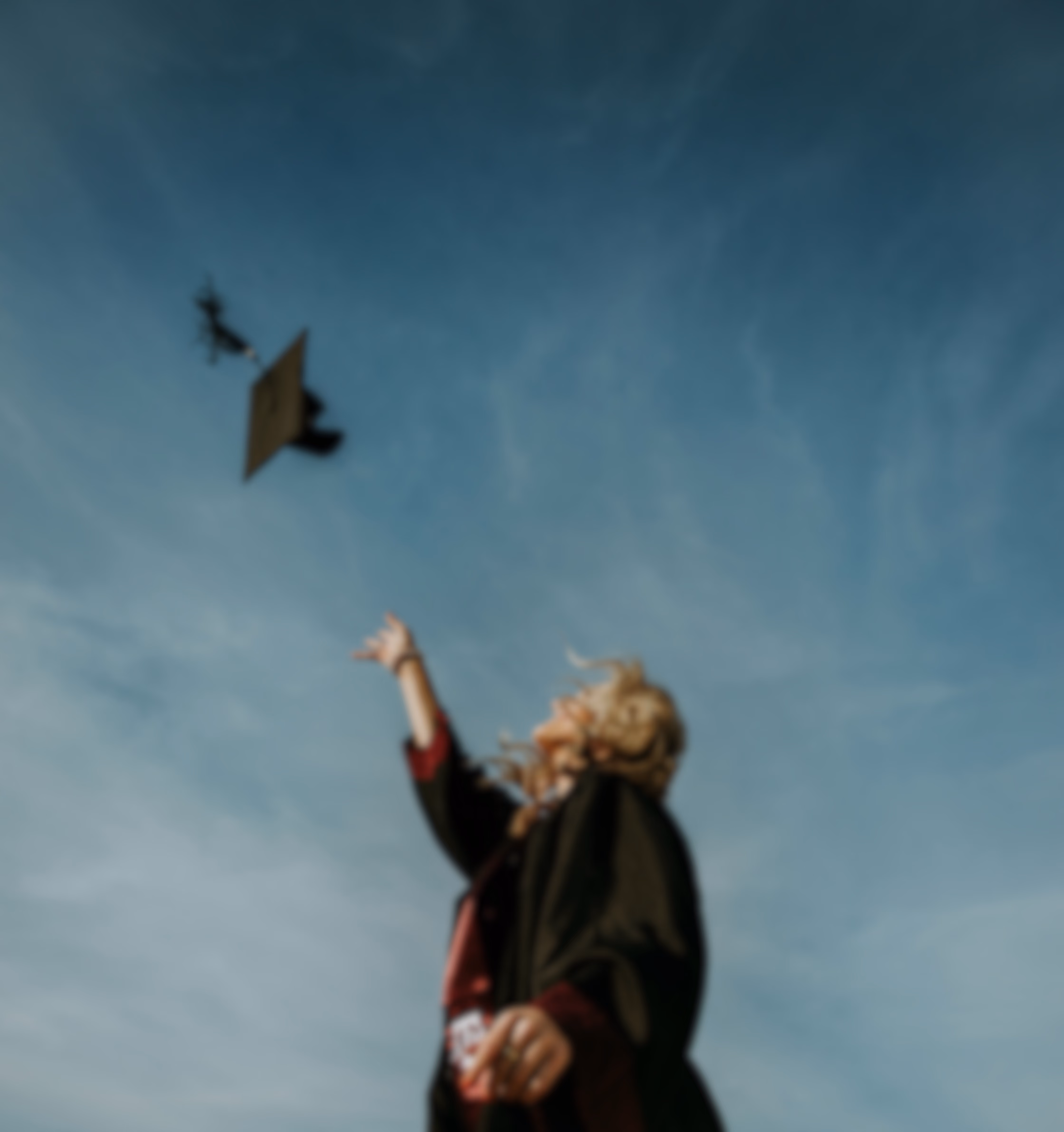 Blurred photo of a graduate in a cap and gown reaching up toward a tossed graduation cap against a blue sky.