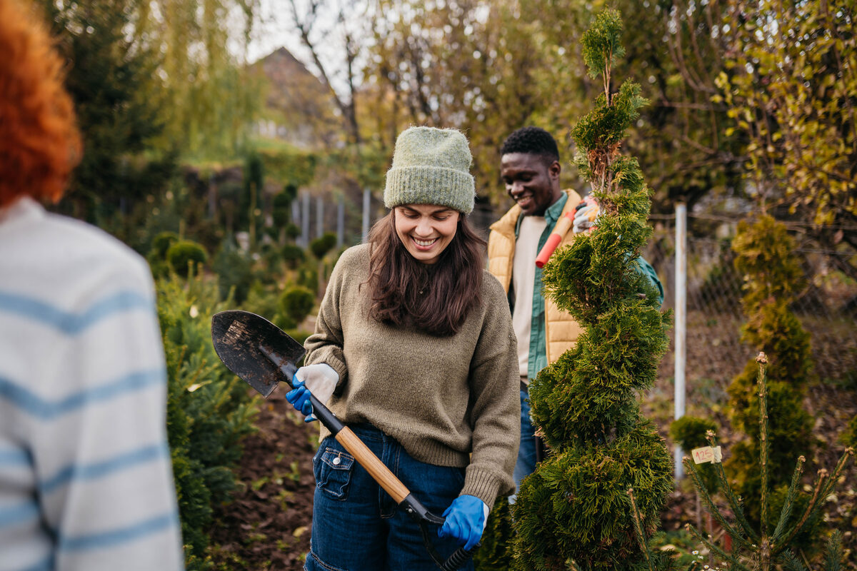 A smiling woman in a green knit hat and blue gloves holds a shovel while a man behind her carries a pruning tool among spiral-shaped green bushes in an outdoor garden center.