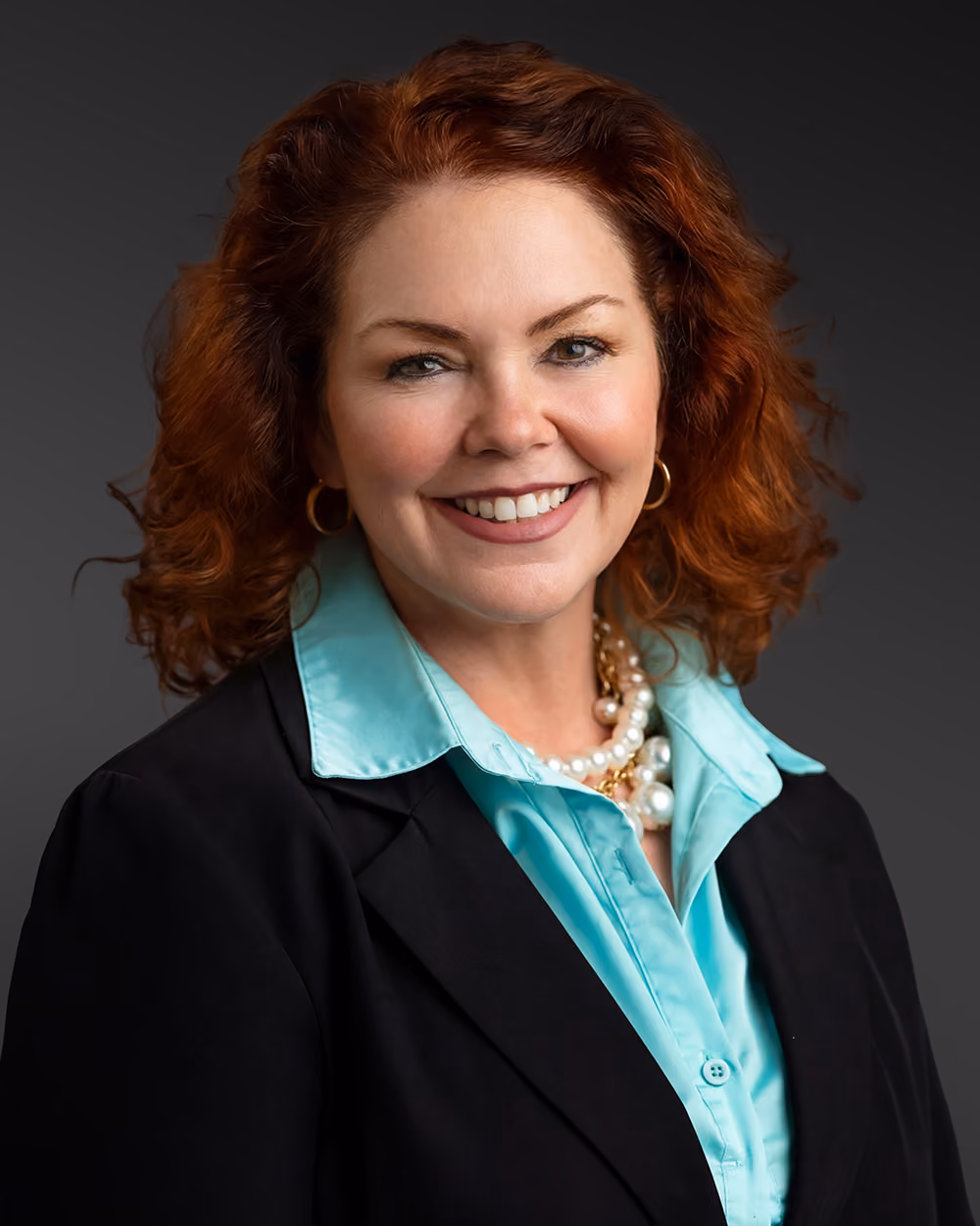 Smiling woman with curly red hair wearing a black blazer, turquoise blouse, and pearl necklace.