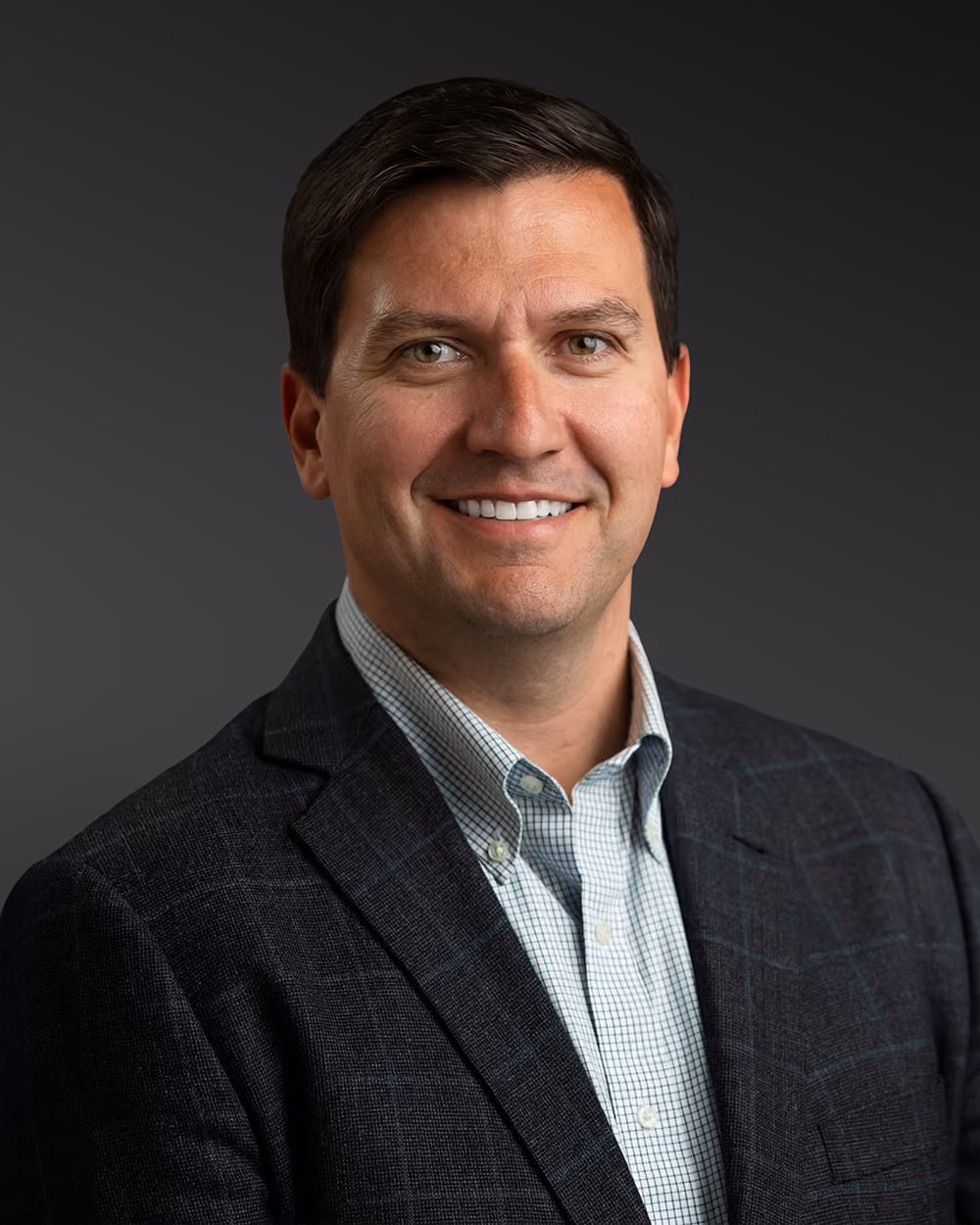 Smiling man with dark hair wearing a checkered shirt and dark blazer against a plain dark background.