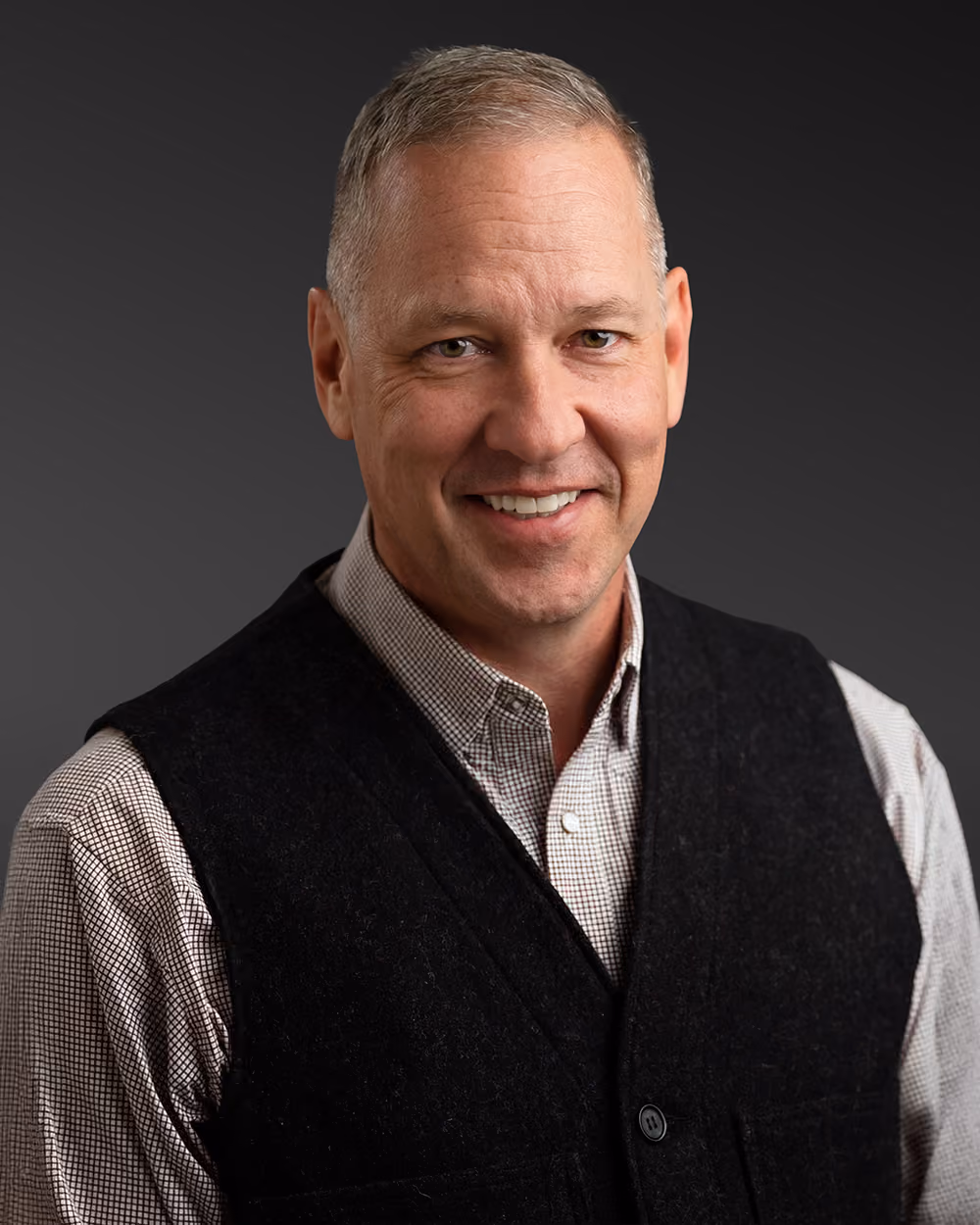 Smiling middle-aged man with short gray hair wearing a checkered shirt and black vest on dark background.